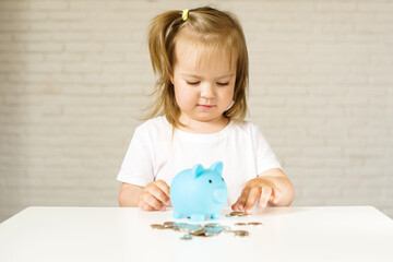 Little Caucasian girl putting coins in to the pig-shaped money box, blue piggy bank. The child manages and deposits his finance, girl saving money in a piggybank for future. 