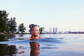 A beautiful smiling woman girl swimming in sea in helsinki Kalasatama behind her summer during evening twilight sunlight sunset in nordic green city travel tips - obrazy, fototapety, plakaty