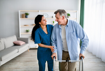 Female caregiver assisting elderly man to walk with frame at home. Professional health care service...