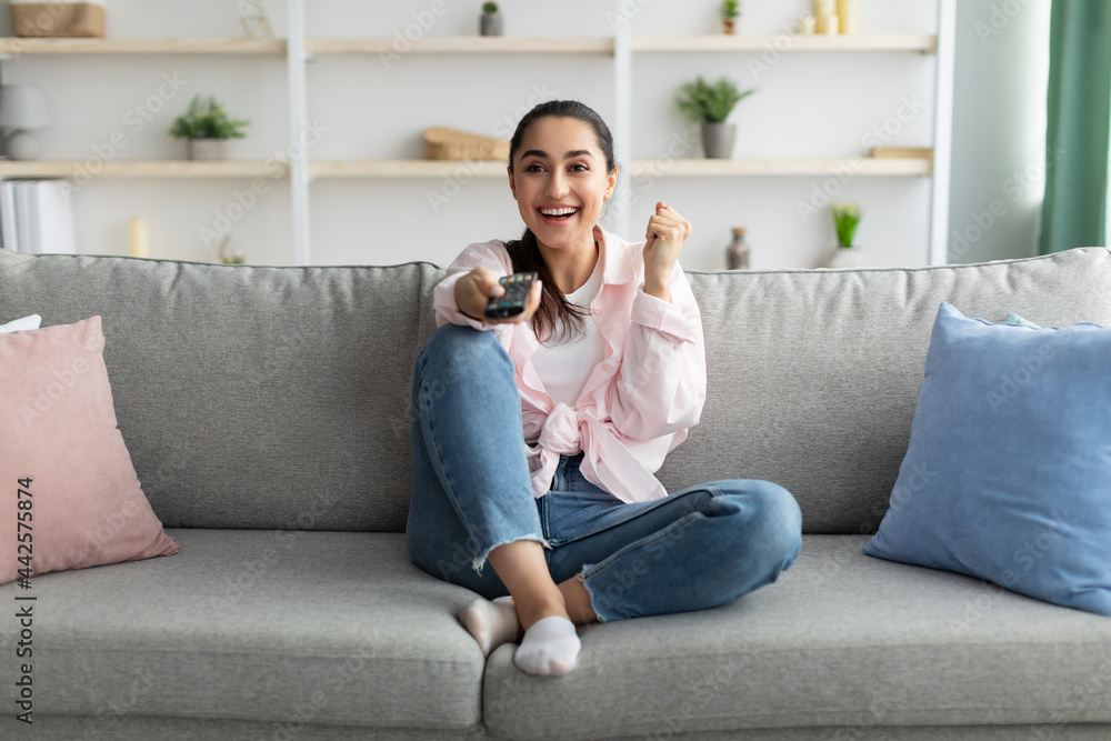 Wall mural Excited woman watching tv shaking clenched fist holding remote control