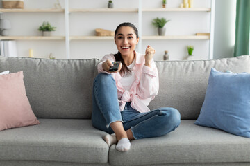 Excited woman watching tv shaking clenched fist holding remote control