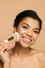 smiling african american woman applying face powder with cosmetic brush isolated on beige