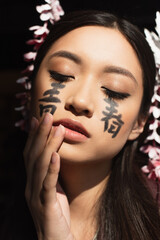 Japanese woman with hieroglyphs on face standing with closed eyes isolated on black