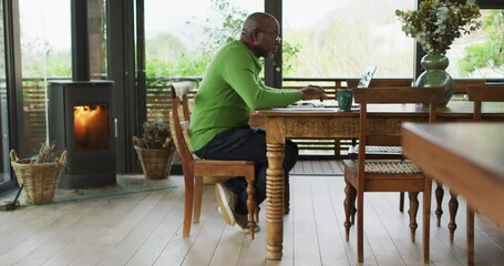 African american senior man sitting at dining table working, using laptop