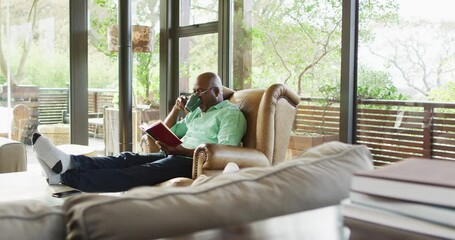 Happy african american senior man relaxing in armchair, feet up, reading book and drinking coffee - Powered by Adobe