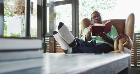 Happy african american senior man relaxing in armchair with feet up, reading a book