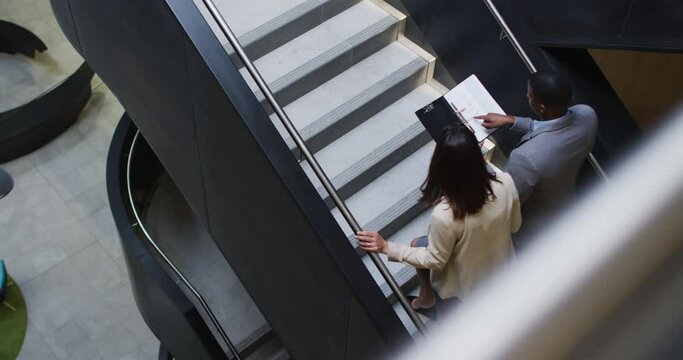 Diverse businessman and businesswoman talking and talking down stairs of modern office