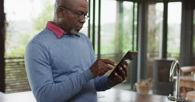 African american senior man at counter in kitchen using tablet, looking away