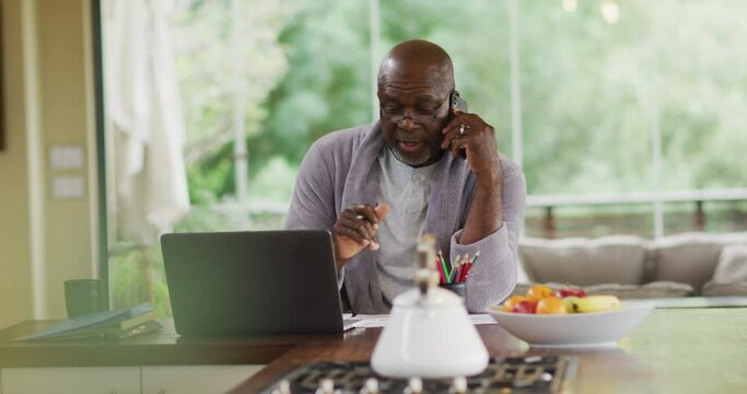 African american senior man in bathrobe sitting in kitchen using laptop and talking on smartphone