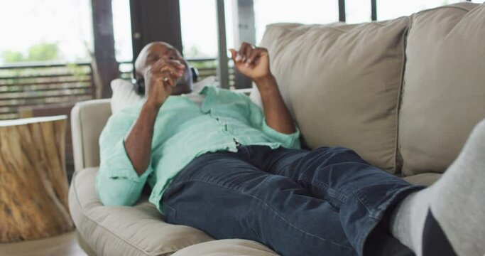 Happy african american senior man relaxing, lying on couch wearing headphones and listening to music