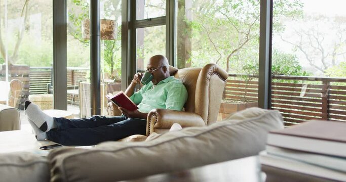 Happy african american senior man relaxing in armchair, feet up, reading book and drinking coffee