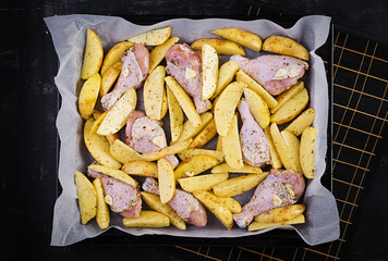 Raw chicken drumsticks and potato wedges in spices on a baking sheet prepared for baking. Top view, flat lay