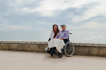 Cheerful adult daughter and aged father in wheelchair on promenade