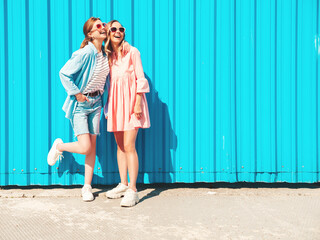 Naklejka premium Two young beautiful smiling hipster female in trendy summer clothes and dress.Sexy carefree women posing in the street near blue wall.Positive pure models having fun at sunset, hugging and going crazy
