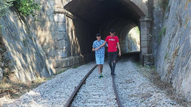 Pareja gay caminado de frente y alegremente por los railes del tren a la salida de un t&uacute;nel del tren haciendo equilibrio sobre el rail.