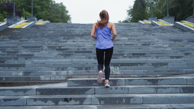 Fit woman with ponytail wearing sports top and leggings jogging on wet staircase in park in slow motion. Following shot athletic female training outside after rain. Concept of fitness