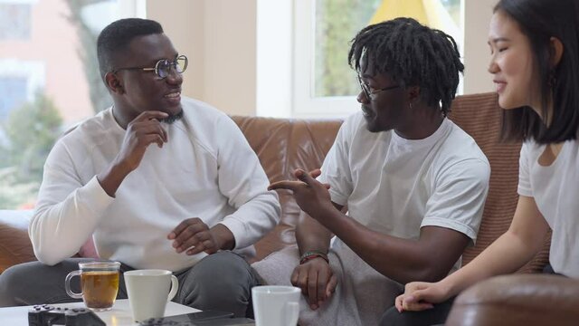 African American Millennial Men Talking Gesturing In Slow Motion Smiling Sitting With Asian Woman Indoors At Home. Positive Happy Relaxed Multiracial Roommates Resting Together Indoors