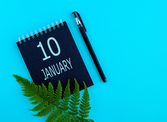 January 10th. Day 10 of month, Calendar date. Black notepad sheet, pen, fern twig, on a blue background. Winter month, day of the year concept