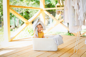 cute caucasian toddler girl in a basket with clean linen on the veranda, hangs up the washed, and...