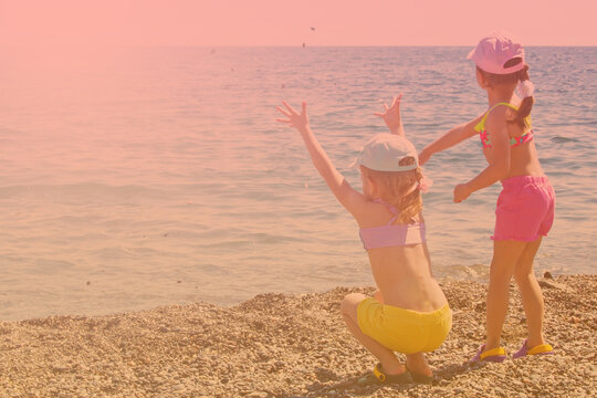 Two girls of five years old in caps, swimsuits, shorts and flip-flops are merrily throwing stones into the sea. Back view. Children on the right side of the frame. The image is toned. Copy space.
