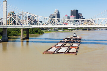A tugboat pushing barges full of sand and gravel down the Ohio River under a bridge in downtown...