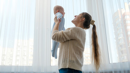 Young mother holding her little baby son against big window at house. Concept of family happiness and child development