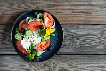 Appetizing salad with feta cheese, fresh vegetables (tomatoes, cucumbers, onions), crispy lettuce, microgreens and violet flowers.  Greek salad on a wooden background. Food with flowers