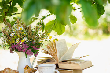 Bouquet of meadow flowers, croissant, cup of tea or coffee, books on table in summer idyllic garden. Rest in garden, reading books, breakfast, vacations in nature concept. Summertime in garden 