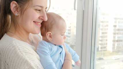 Portrait of smiling mother with little baby son looking on city street through window. Concept of family happiness and child development