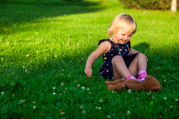 Cute little blond baby girl two year old playing with teddy bear on fresh green grass with flowers. Kid having fun making first steps on mowed natural lawn. Happy and healthy childhood concept