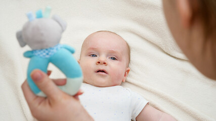 Young mother talking to her little baby in bed and shaking colorful rattle toy. Concept of baby education and development.