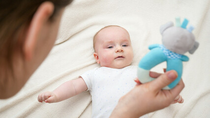 Cute little baby lying in bed and looking on mother and colorful rattle toy. Concept of baby education and development.