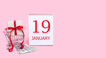 A gift box in a shopping trolley, dollars and a calendar with the date of 19 january on a pink background.