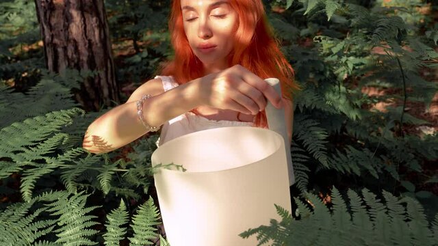 Red-haired woman playing on white quartz crystal singing bowls in the forest. Healing medicine and meditation Spiritual music Buddha Fern background