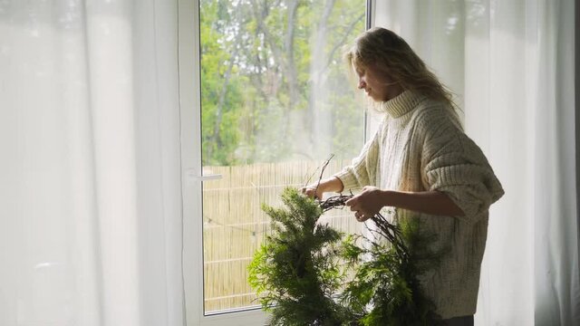 Young blondie woman in white sweater makes festive wreath for Christmas holiday with thuja twigs while standing near large window with curtains at home