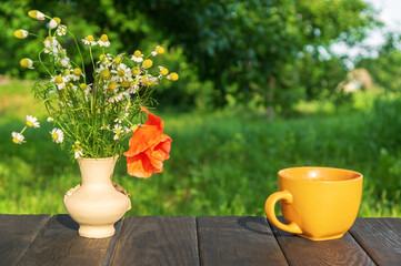 Yellow cup of hot coffee or tea and a bouquet of flowers on a wooden table in a green garden in the morning or evening. Summer mood