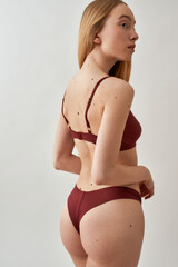 Back view of attractive young slim caucasian woman with beautiful perfect body wearing burgundy underwear looking aside, posing isolated over light gray background