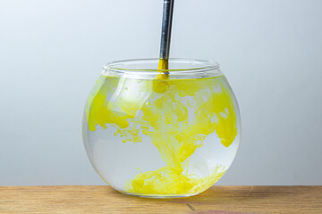A paintbrush with yellow paint is dipped in a jar of water on a white background. The paint spreads in the water.