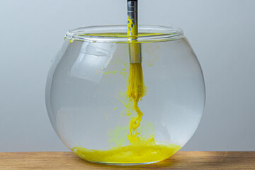 A paintbrush with yellow paint is dipped in a jar of water on a white background. The paint spreads in the water.