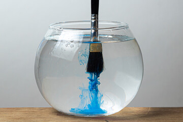 A brush with blue paint is dipped in a jar of water on a white background. The paint spreads in the water.