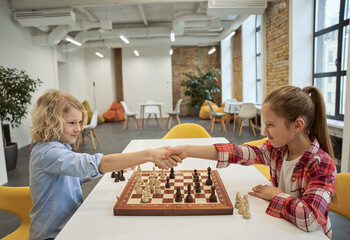 Rivalry. Two little children, boy and girl shaking hands after match, playing board game, sitting together at the table in school