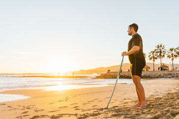 Caucasian hispanic man standing on the beach at sunset with a skimboard clutched in his hands - copyspace