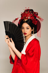 Pretty asian woman in red kimono holding fans and looking at camera isolated on grey