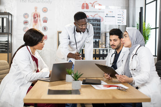 Four Diverse Medical Workers In Uniform Having Discussion At Office With Modern Gadgets On Table. Health Care And Teamwork Concept.