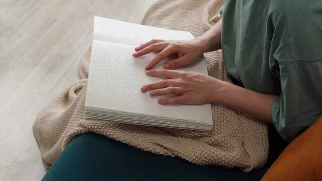 Blind woman reading braille book sitting on sofa, touching letters on sheet of paper using his fingers, poorly seeing person learning to read, concept of home education for people with disabilities.
