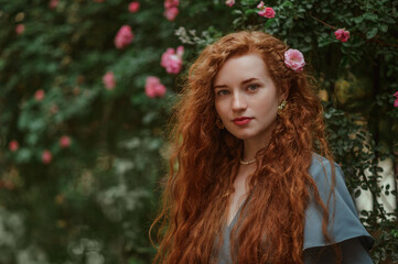 Beautiful redhead freckled woman with long curly natural hair, posing outdoor, near blooming roses....