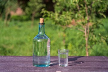 Glass and big bottle with drinking water on a wooden table on nature background in garden. It can be water or vodka