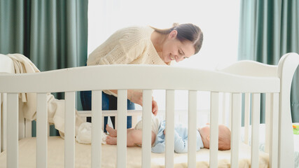 Beautiful smiling mother undressing her little baby son lying in cradle at morning to change diapers. Concept of parenting, family happiness and baby development.