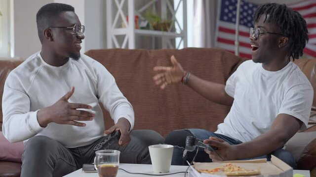 Positive millennial African American men giving high-five rejoicing victory in multiplayer video game. Happy laughing handsome friends winning resting on weekend indoors