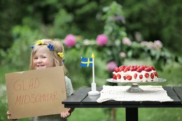 Glad midsommar / Happy Midsummer! Girl holding sign saying "glad midsommar" meaning "happy midsummer" in swedish. Traditional strawberry cake on the side and a swedish flag in the middle.